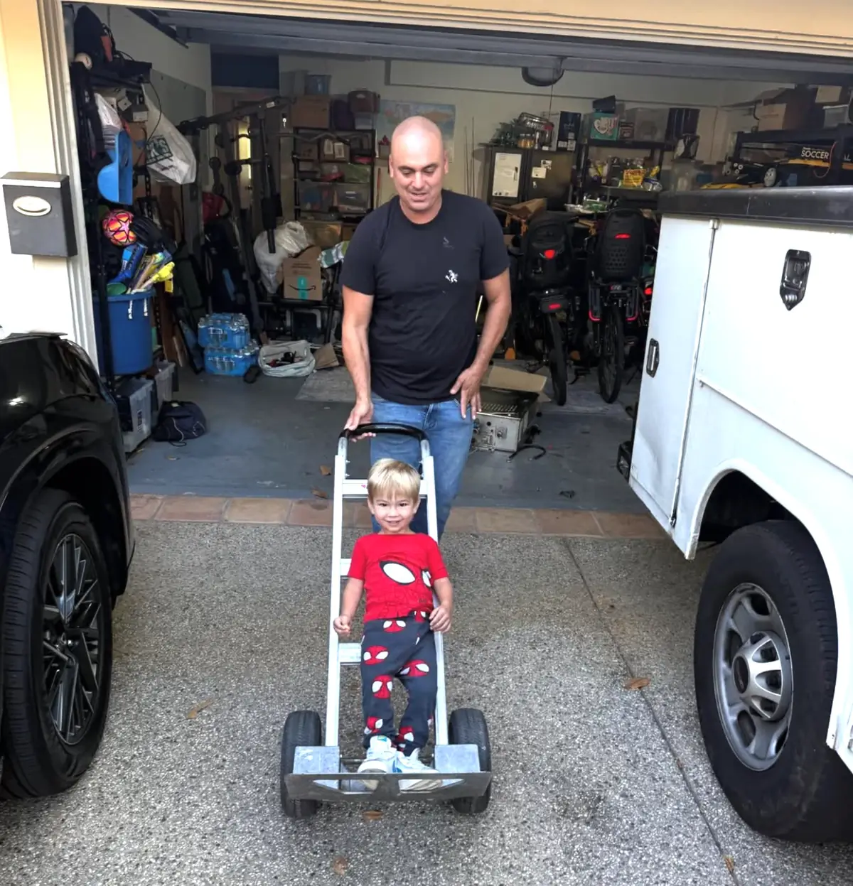 Dan in a driveway pushing a hand truck with a smiling little helper riding on it.