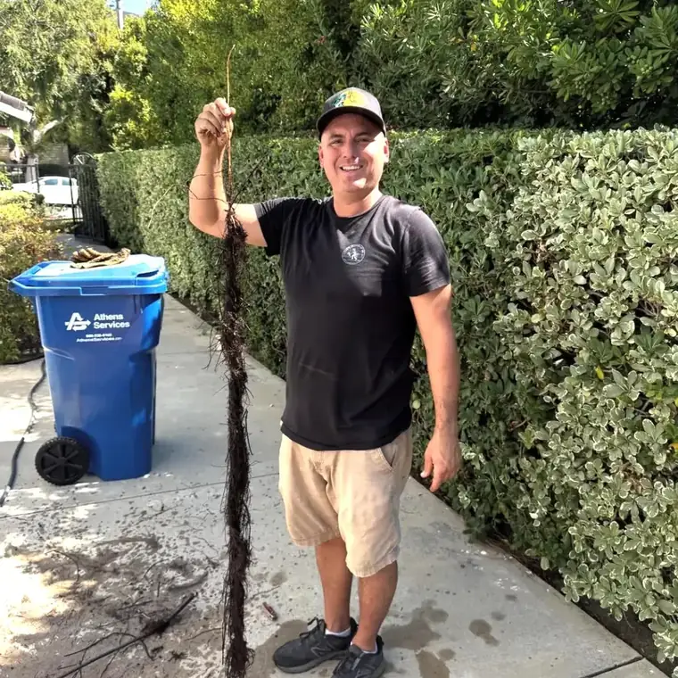 Dan of Dan's Plumbing on a sidewalk by a hedge, smiling and holding up a long clump of tree roots cleared out of a pipe.